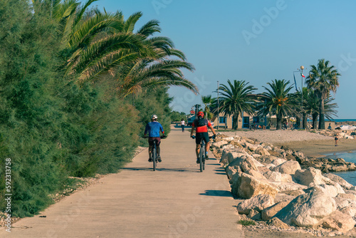 Authentic cyclists ride along the path along the seashore on a sunny summer morning in Italy against a beautiful background of green palms and blue sky, view from the back, healthy lifestyle