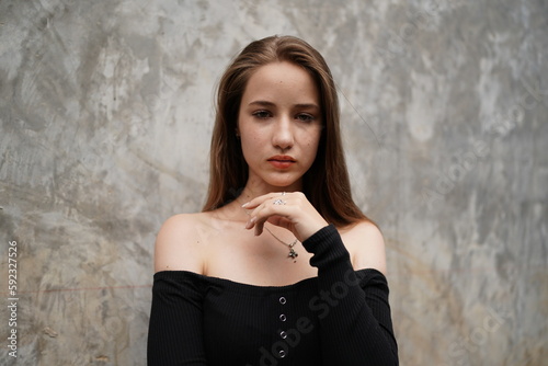 Fotografie Melancholy Gaze: A young woman with long, auburn hair and a pensive expression stands against a textured concrete wall, her eyes conveying a sense of introspection and sadness