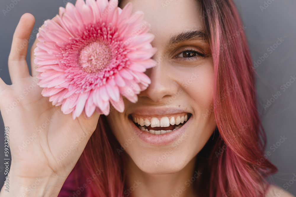 Close up shot of cheerful smiling attractive young pink hair woman covering a part of her face with pink gerbera flower, isolated over grey background. Women cosmetics and face skin care. Happy girl