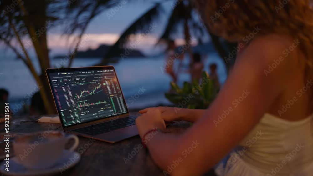 Female cryptocurrency trader at laptop checks charts online working remotely at outdoor tropical seaside cafe at sunset. Woman crypto broker analyses graphic of stock exchange rates by ocean close-up