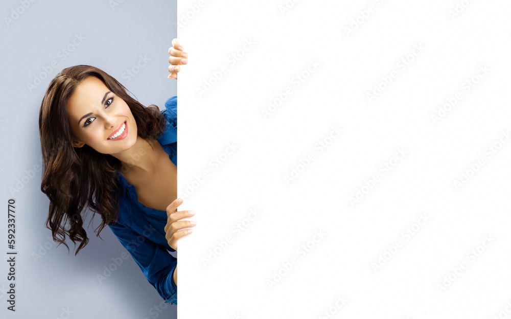 Happy smiling brunette young woman in blue clothing, standing behind, peeping from blank banner or mock up signboard with copy space for ad text, on grey gray background. Happy smiling brunette young woman in blue clothing, standing behind, peeping from blank banner or mock up signboard with copy space for ad text, on grey gray background.