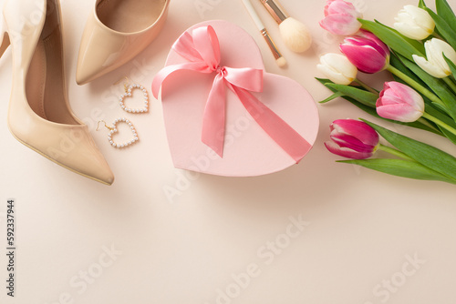 Fashionable Mother's Day concept. Top view flat lay of high-heels, gift box, tulip flowers, makeup brushes, and earrings on a pastel beige background with space for text or advert
