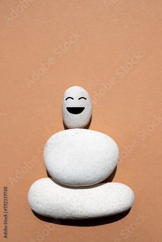 Three white smooth stones on top of each other on a beige background. Stone with a funny face. The concept of balance and positive thinking. Top view, space for text. Vertical image.