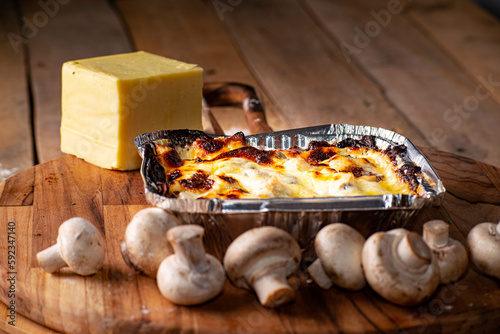 Lasagna or pasta gratin baked in an aluminum tray surrounded by mushrooms and mozzarella cheese on a rustic wooden table.