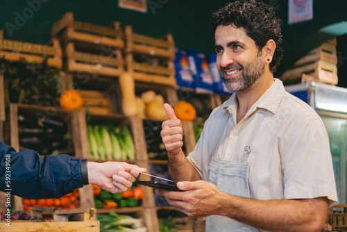 Middle-aged Latin grocer charging by credit card with chip through his cell phone and giving a thumbs up.