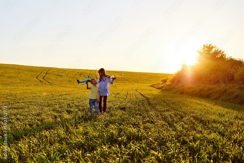Fototapeta premium Running boy and girl holding two yellow and blue airplanes toy in the field during summer sunset. Kids dreams of flying and aviation.