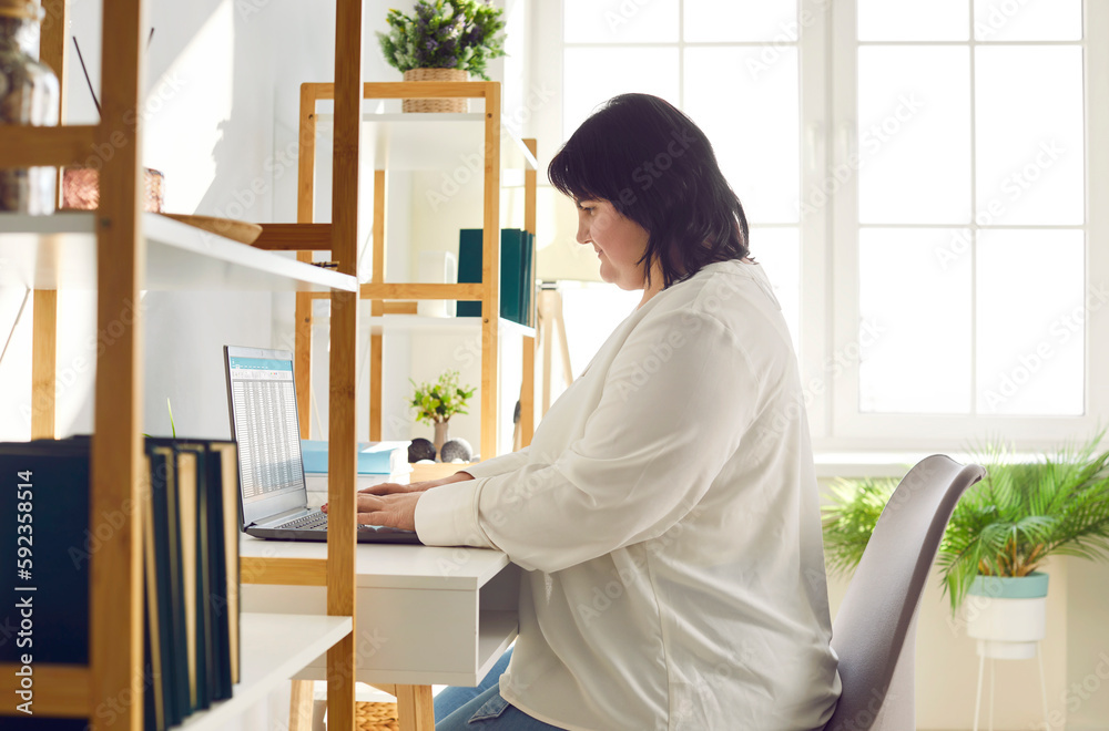 Overweight brunette woman working with laptop in office. Side view shot of female accountant, financial advisor sitting at her desk analyzing financial data on computer screen Overweight brunette woman working with laptop in office. Side view shot of female accountant, financial advisor sitting at her desk analyzing financial data on computer screen