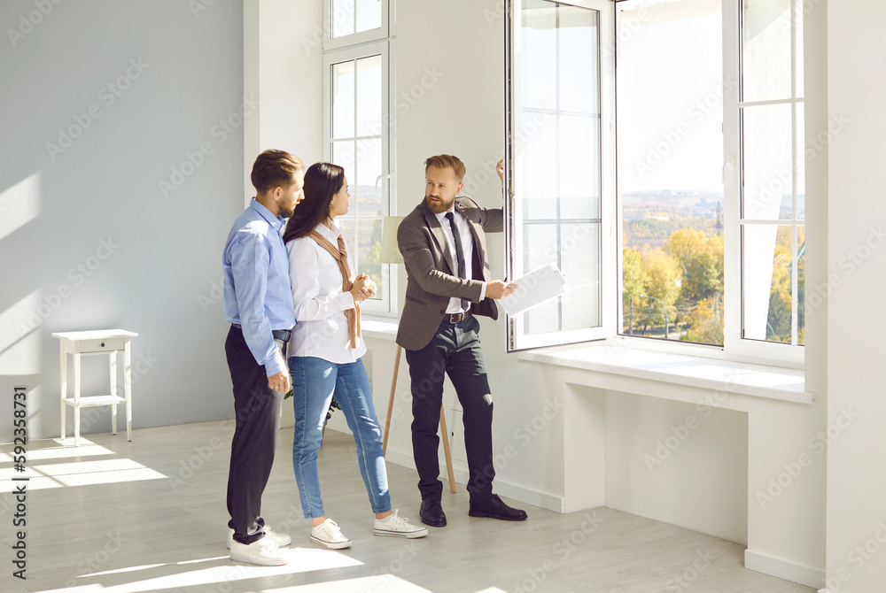 Handsome bearded male realtor in suit with documents in his hands advises young couple before buying new house, demonstrates view from the window. Three people are standing in modern sunny apartment. Handsome bearded male realtor in suit with documents in his hands advises young couple before buying new house, demonstrates view from the window. Three people are standing in modern sunny apartment.