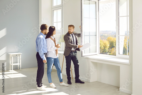 Handsome bearded male realtor in suit with documents in his hands advises young couple before buying new house, demonstrates view from the window. Three people are standing in modern sunny apartment. photo