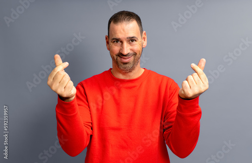Canvastavla Bearded hispanic man in his 40s wearing a red sweater making the money gesture rubbing his fingers with a greedy face, isolated over gray background