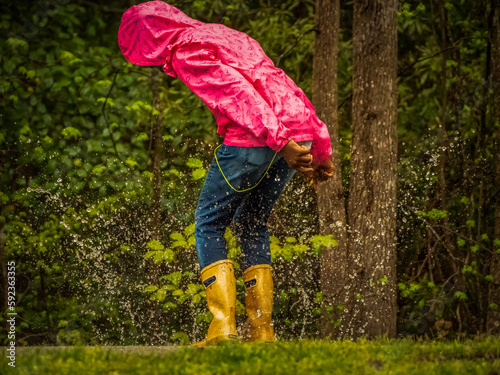 Child in bright pink parka and tan rain boots, looking down and splashing in rain puddle