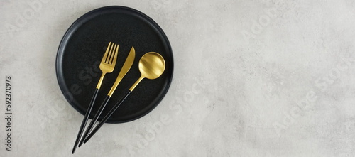 Empty black ceramic plate and cutlery on gray stone background top view with copy space banner . Modern kitchen utensils.Space for text or menu .Business food brand template.