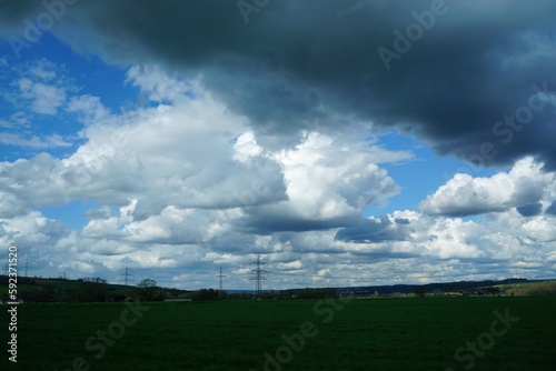Grüne Felder mit Strommasten vor blauem Himmel mit grauweißem großen Wolkengebilde am Nachmittag im Frühling © Anette