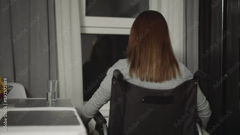 Woman with spinal cord injury looks out window pulling back curtain. Female person peers at cause of flashing beacons lights on street in evening backside view