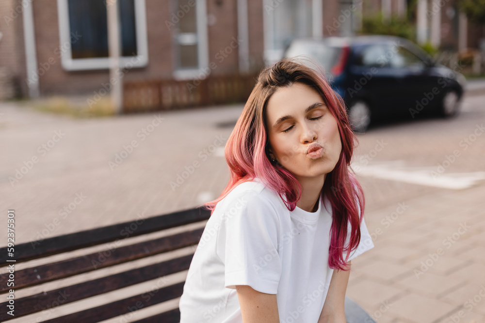 Pensive pink hair woman sitting on bench and make blow kisses walking on the street. Outdoor shot of happy hippie lady in boho freedom style. Girl send air kiss to camera, closed eyes, love concept.