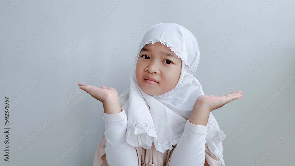 A portrait of an Asian child wearing hijab looks so confused between choices, isolated on gray background.