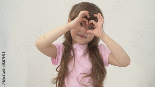 A portrait happy little girl child with brown hair smiles and holding heart shaped her hands on eyes. Health of eyes and vision. Copy space. 
