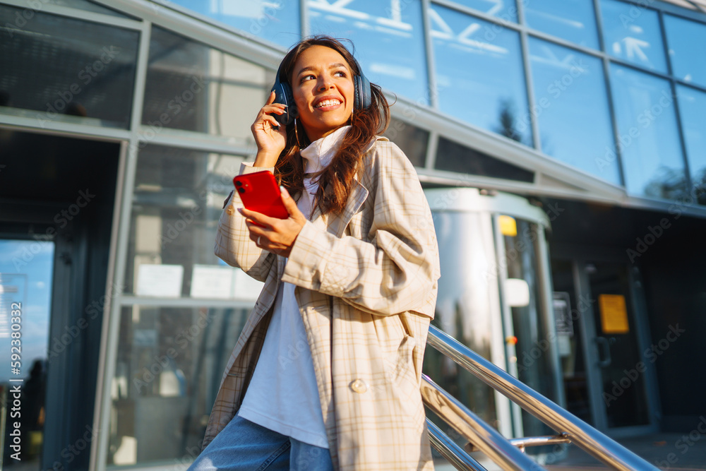 Optimistic woman wearing headphones and holding phone outdoors. Young woman listens to music, audiobook or podcast while walking on sunny street. Communication in social networks. Lifestyle.