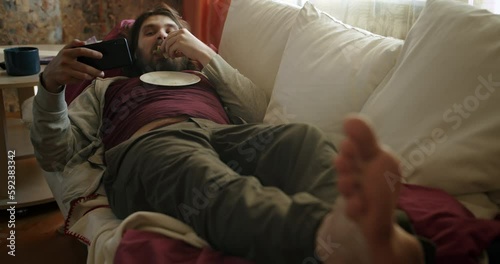 Carefree, careless lazy beard man eating sandwich, watching some entertaining content on his smartphone, laughing, lying on the couch. Eating disorder, overweight. Laziness, procrastination concept.