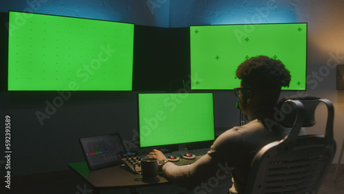 African American man works on computer in studio with equipment. Multiple chroma key big screen on the wall. Professional software displayed on tablet. Color grading or film editing room. Dolly shot.