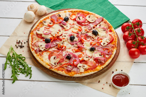 Freshly baked pizza with smoked sausages, red pepper, mushrooms, onion and olives served on wooden background with tomatoes, sauce and herbs. Food delivery concept. Restaurant menu