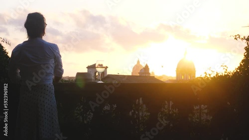 Young woman tourist fashion white dress with glass of white wine in front of panoramic view of Rome cityscape from campidoglio terrace at sunset. Landmarks, domes of Rome, Italy.