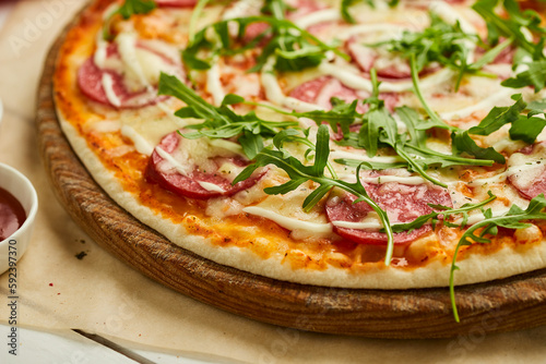 Freshly baked tasty pepperoni pizza with salami, mozzarella cheese and rukkola served on wooden background with tomatoes, sauce and herbs. Food delivery concept. Restaurant menu