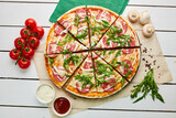 Sliced Freshly baked tasty pepperoni pizza with salami, mozzarella cheese and rukkola served on wooden background with tomatoes, sauce and herbs. Food delivery concept. Restaurant menu