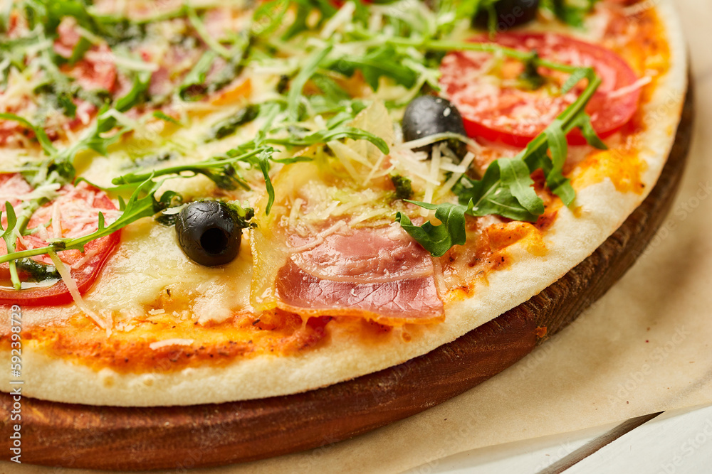 Freshly baked pizza with ham, rukkola, sauce pesto and olives served on wooden background with tomatoes, sauces and herbs. Food delivery concept. Restaurant menu