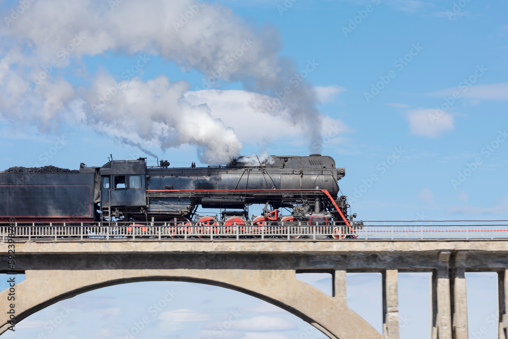 A smoking steam locomotive driving over an old bridge against a blue sky