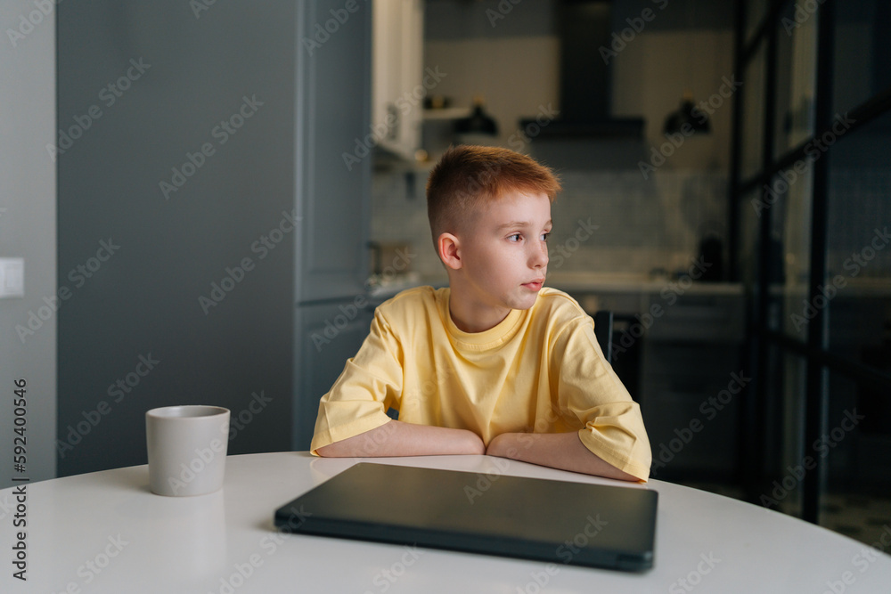 Portrait of pensive redhaired 10 year old boy sitting in front of closed laptop, thoughtful looking away. Front view of cute child boy using computer sitting at table in modern kitchen room. Portrait of pensive redhaired 10 year old boy sitting in front of closed laptop, thoughtful looking away. Front view of cute child boy using computer sitting at table in modern kitchen room.