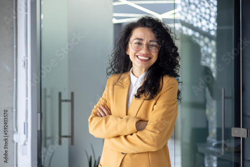 Portrait of successful business woman inside office, latin american boss in yellow suit smiling and looking at camera, mature female worker with curly hair standing with arms crossed.