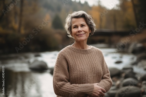 Environmental portrait photography of a pleased woman in her 50s wearing a cozy sweater against a peaceful river or stream background. Generative AI