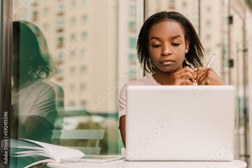Serious calm face african american woman hipster sitting outside street cafe with laptop, working typing message. Young entrepreneur female freelancer high school student businesswoman
