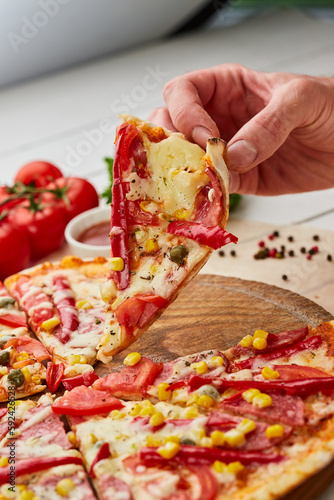 The human hand takes Freshly baked tasty pepperoni pizza with salami, mozzarella cheese, corn and pepper served on wooden background with tomatoes, sauce and herbs. Food delivery concept