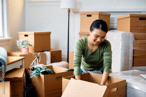 Happy Asian woman packing her belongings in cardboard boxes while relocating in new apartment.