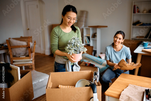 Happy Asian woman and her female roommate unpacking cardboard boxes while moving into new apartment.