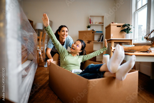 Cheerful woman has fun while friend is pushing her in cardboard box at their new home. photo