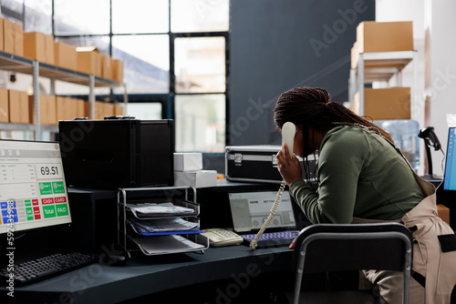 African american manager talking at landline phone with remote customer, discussing order details in storage room. Warehouse employee wearing industrial overall while working at goods inventory