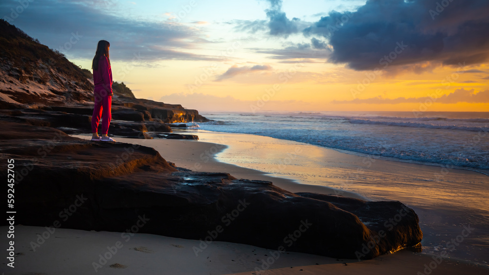 A beautiful girl in a pink tracksuit stands on a rock on Ten Mile Beach and enjoys a stunning orange sunrise over the Pacific ocean. Black rocks campsite, NSW, Australia A beautiful girl in a pink tracksuit stands on a rock on Ten Mile Beach and enjoys a stunning orange sunrise over the Pacific ocean. Black rocks campsite, NSW, Australia