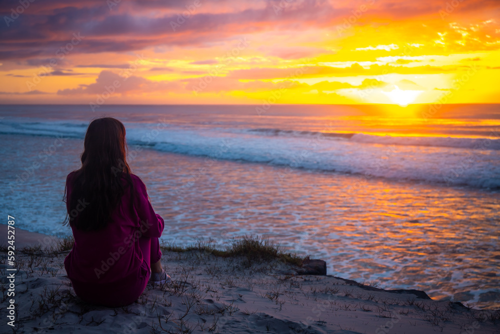Girl in pink tracksuit sits on magnificent Ten Mile beach and enjoys colorful sunrise over Pacific ocean. Black rocks campground, NSW, Australia