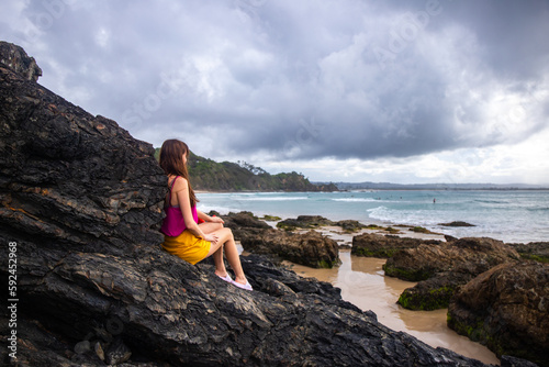 Beautiful girl stand on large rocks and admires powerful  waves in popular Byron Bay, NSW, Australia
