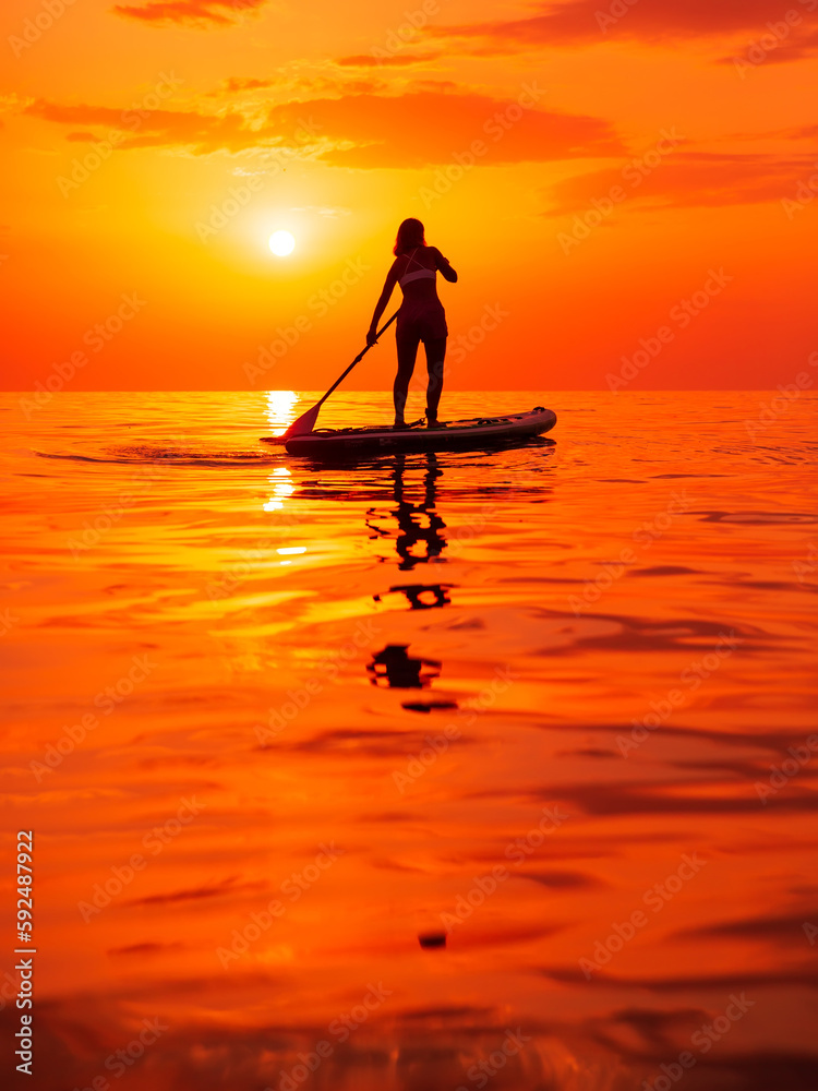 Silhouette of woman rowing on stand up paddle board at sea with warm sunset or sunrise.