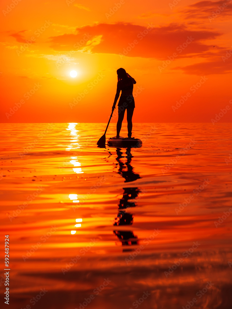 Silhouette of woman on stand up paddle board at sea with warm sunset or sunrise. Woman rowing on SUP board and bright sunset with reflection on water