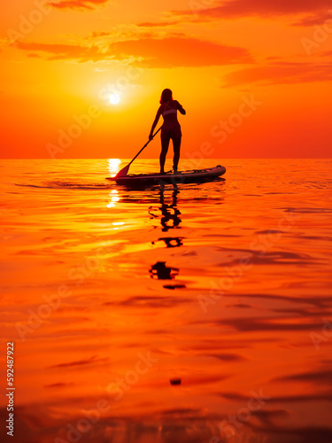 Silhouette of woman rowing on stand up paddle board at sea with warm sunset or sunrise.
