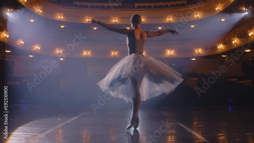 Ballet female dancer in white tutu dress practices choreography moves on stage and prepares to start a show. Ballerina on rehearsal of performance. Illuminated theatrical hall. Classical ballet art.