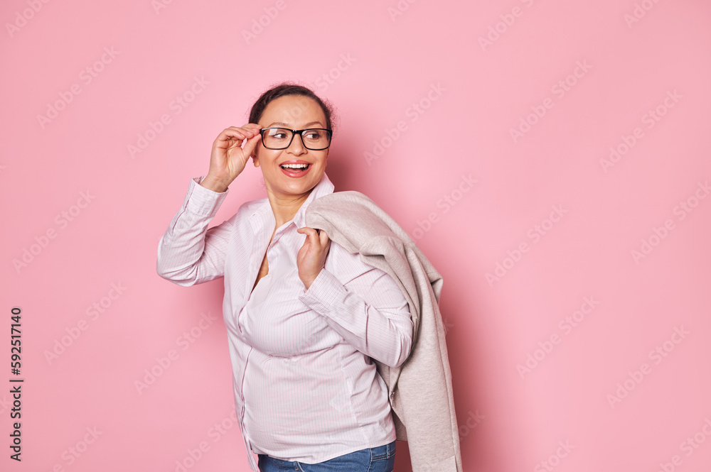Pregnant cheerful ethnic woman smiling with beautiful toothy smile, looking aside, putting trendy glasses on isolated pink background. Copy ad space. Business. People. Pregnancy. Maternity leave