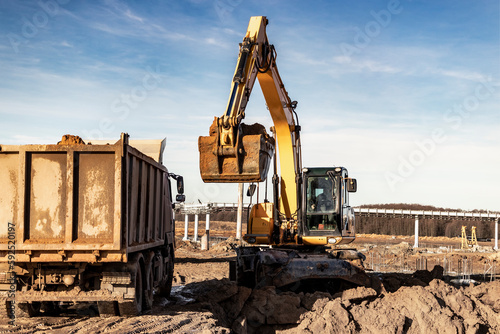 A wheeled excavator loads a dump truck with soil and sand. An excavator with a high-raised bucket against a cloudy sky View from the trench. Removal of soil from a construction site or quarry.