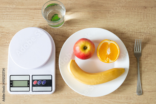 An apple orange and a banana on a plate in the form of a smiling person, next to a kitchen scale and a glass of water, a close-up view from above. Proper nutrition