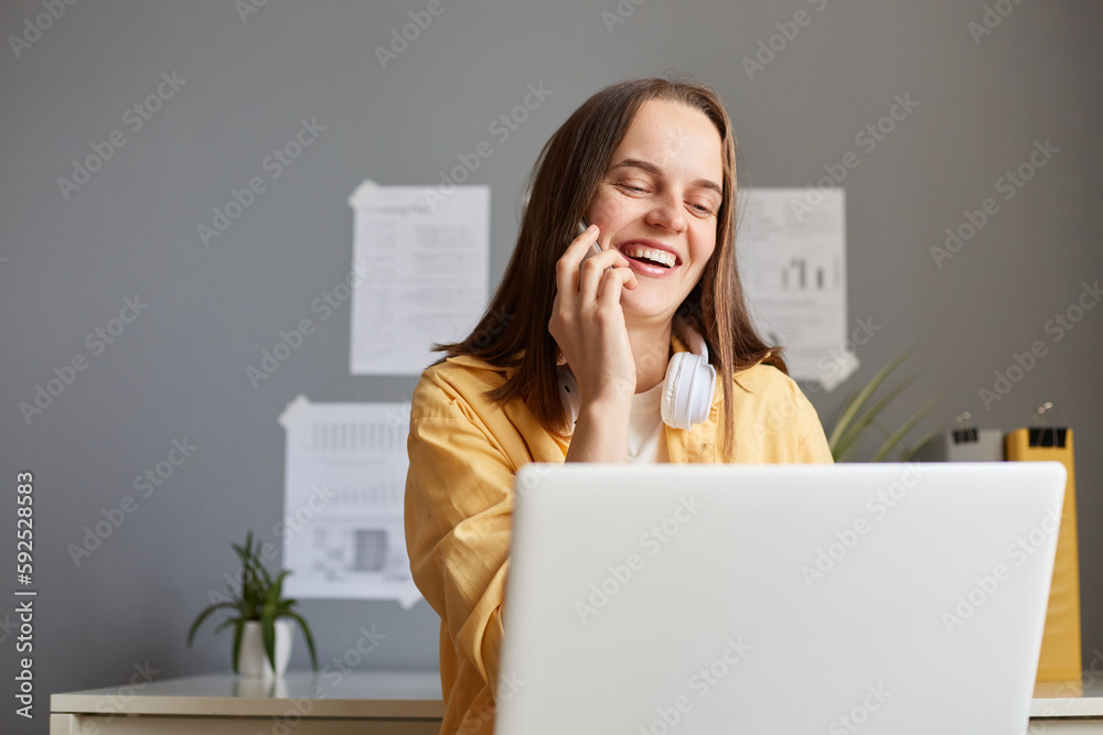 Smiling happy hipster woman working on laptop at office while talking on phone, having break, talking with friend, laughing, resting, having funny conversation. Smiling happy hipster woman working on laptop at office while talking on phone, having break, talking with friend, laughing, resting, having funny conversation.